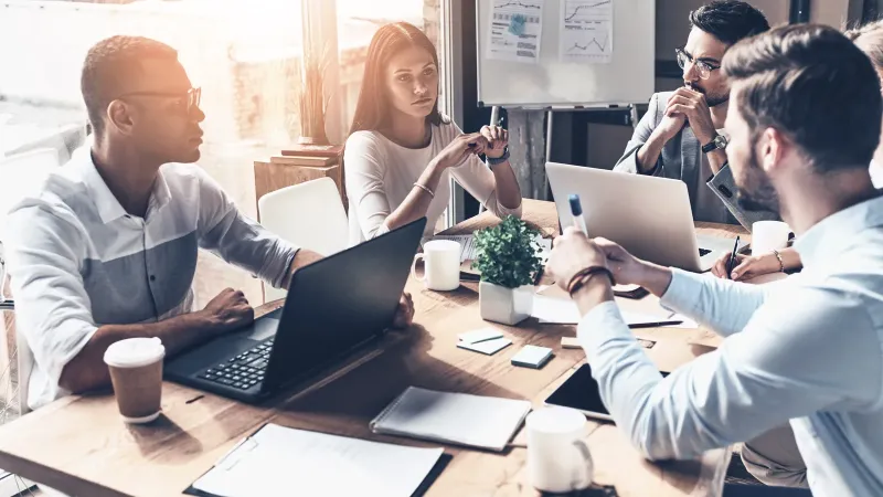 Cuatro profesionales conversan alrededor de una mesa de reuniones con portátiles y un tablero con gráficas al fondo