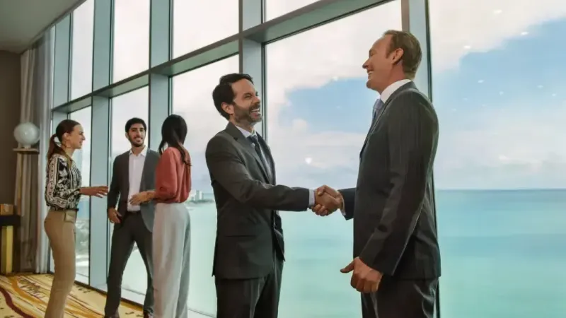 Dos ejecutivos se dan la mano en una sala con ventanal sobre el mar, mientras otros colegas conversan al fondo