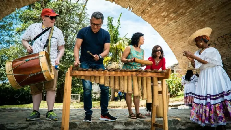 Visitantes tocando marimba y tambor con músicos tradicionales del Pacífico bajo un puente de ladrillo